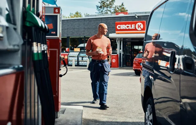 Man happily returning to his vehicle at a Circle K station with a sandwich and coffee in hand